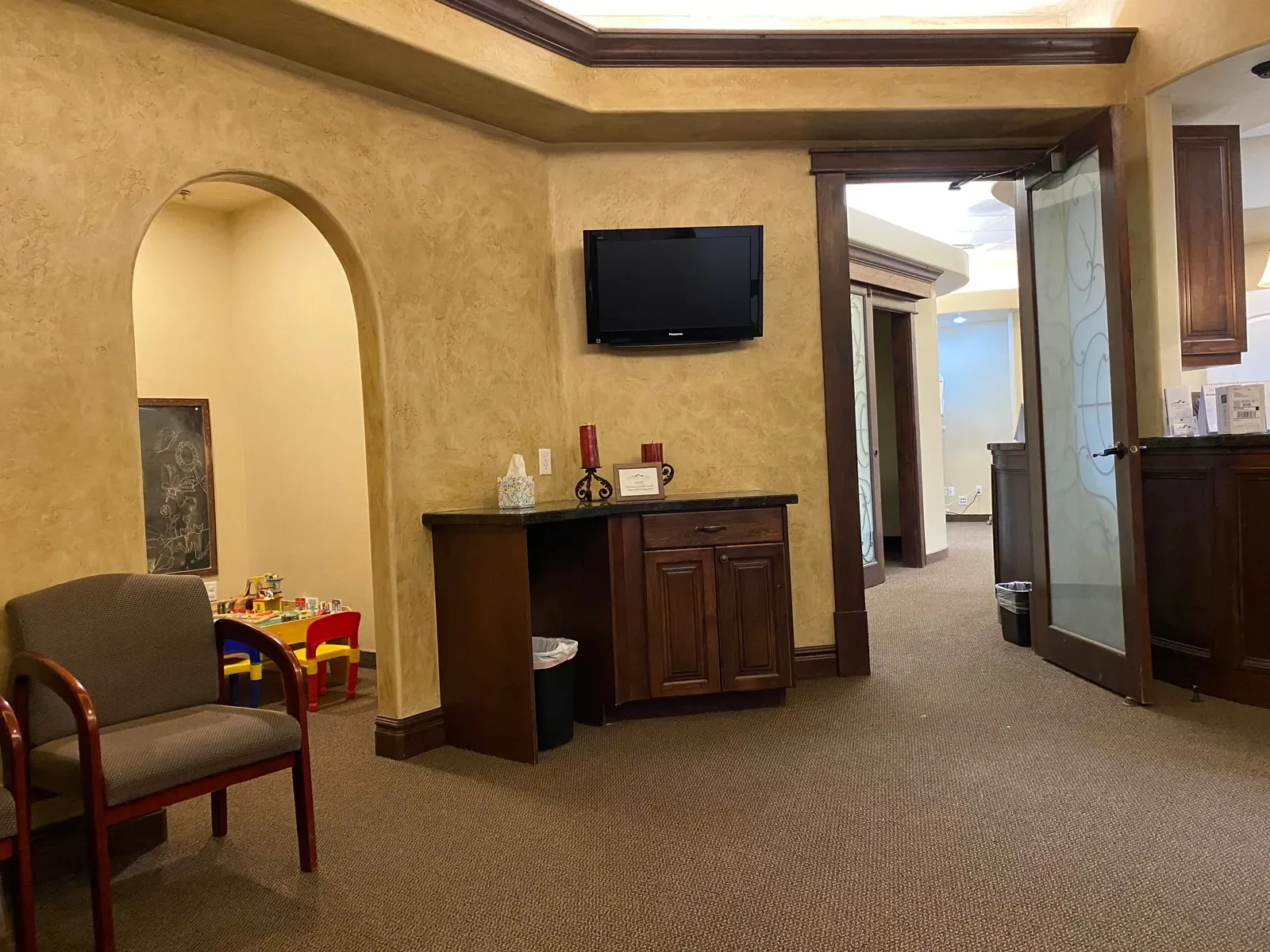 Waiting room with an armchair, wall-mounted TV, and an arched entryway leading to a children's play area.